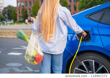 Woman holding a mesh bag with vegetables and charging an electric car in the street, back view. 118901372