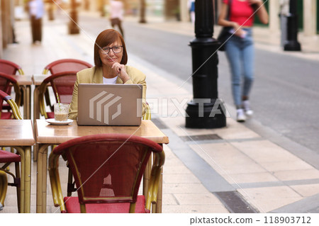 Mature stylish confident businesswoman in eyeglasses and suit works on laptop drinks coffee at European cafe outdoors. Concept of remote work from public place, digital freelance and modern lifestyle Mature stylish confident businesswoman in eyeglasses and suit works on laptop drinks coffee at European cafe outdoors. Concept of remote work from public place, digital freelance and modern lifestyle 118903712