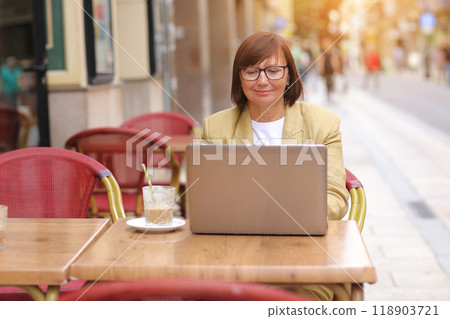 Mature stylish businesswoman in eyeglasses works on laptop while sitting with a coffee drink at European cafe outdoors. Concept of remote work from public place, digital freelance and modern lifestyle 118903721