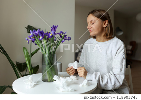 Seasonal allergy free. Young woman sniffs of iris flowers, enjoys of smell without of runny nose, itching or cough seasonal symptoms at cozy home. Girl sits by the table. Used paper tissues on table 118903745