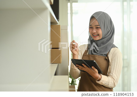 Smiling young woman wearing a hijab using digital tablet managing inventory or orders in a small business Smiling young woman wearing a hijab using digital tablet managing inventory or orders in a small business 118904153
