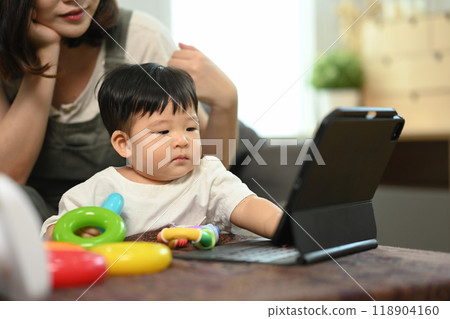 Cute little toddler boy sitting in living room with colorful toy and looking at digital tablet Cute little toddler boy sitting in living room with colorful toy and looking at digital tablet 118904160