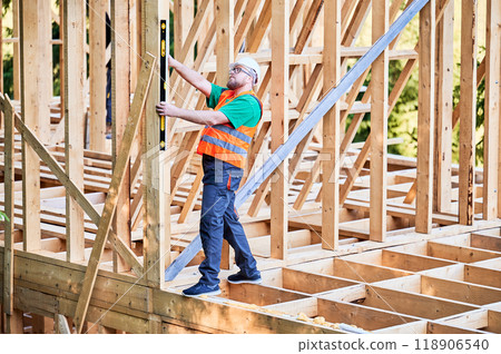 Carpenter constructing two-storey wooden frame house. Man inspects walls for levelness using spirit level, wearing protective overalls, helmet and vest. Concept of modern ecological construction. Carpenter constructing two-storey wooden frame house. Man inspects walls for levelness using spirit level, wearing protective overalls, helmet and vest. Concept of modern ecological construction. 118906540
