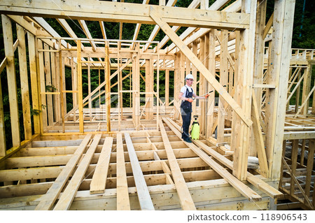 Laborer constructing wooden frame house near forest. Man treating woods, applying fire retardant using sprayer, while dressed in protective suit, helmet. Concept of modern eco-friendly construction. 118906543