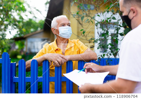 Senior man in protective mask communicating with insurance company representative while standing at fence of his country house 118906704