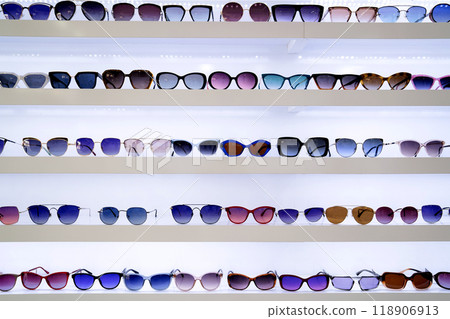 Organized rows of sunglasses, vibrant lenses in different styles, white shelves displaying fashion accessories, retail presentation enhancing shopping ambiance, front view. 118906913