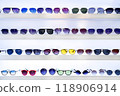 Rows of sunglasses in various styles and colors on white shelves, showcasing eyewear options, vibrant lens hues, fashion accessories, retail store presentation, organized selection, shopping ambiance 118906914