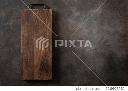 Wooden cutting board with metal handle on dark brown kitchen table top. Space for design. Top view. Cooking concept. Wooden cutting board with metal handle on dark brown kitchen table top. Space for design. Top view. Cooking concept. 118907281