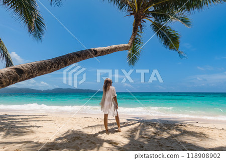 A young woman enjoys a peaceful moment on a tropical beach under a palm tree with clear blue waters in the background on a sunny day A young woman enjoys a peaceful moment on a tropical beach under a palm tree with clear blue waters in the background on a sunny day 118908902