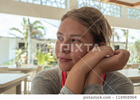A young woman sits at a cafe table, resting her chin on her arms and gazing thoughtfully outside. 118908904