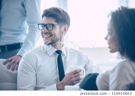 Life of businessman. Close-up part of young handsome cheerful man holding coffee cup and looking away with smile while sitting in office with his coworkers Life of businessman. Close-up part of young handsome cheerful man holding coffee cup and looking away with smile while sitting in office with his coworkers 118910663