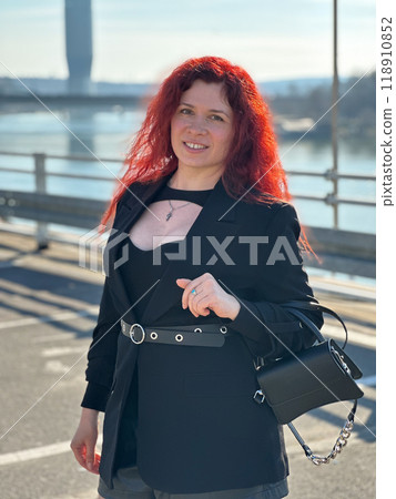 Millennial woman with long curly red hair gazing thoughtfully at the camera on a sunny day. Background features modern buildings and a river, evoking a sense of urban calm. City life, Belgrade and Millennial woman with long curly red hair gazing thoughtfully at the camera on a sunny day. Background features modern buildings and a river, evoking a sense of urban calm. City life, Belgrade and 118910852