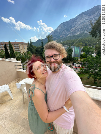 Millennial couple enjoys a vacation with a beautiful mountain backdrop while smiling and embracing on a sunny day taking selfie. Terrace with a scenic view of lush greenery and residential buildings 118911021
