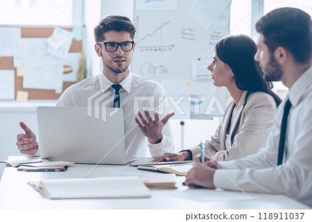 Brainstorming with business partners. Young handsome man in glasses gesturing and discussing something with his coworkers while sitting at the office table Brainstorming with business partners. Young handsome man in glasses gesturing and discussing something with his coworkers while sitting at the office table 118911037