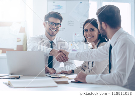 Congratulations! Two handsome man shaking hands with smile while sitting at the office table with their beautiful coworker Congratulations! Two handsome man shaking hands with smile while sitting at the office table with their beautiful coworker 118911038