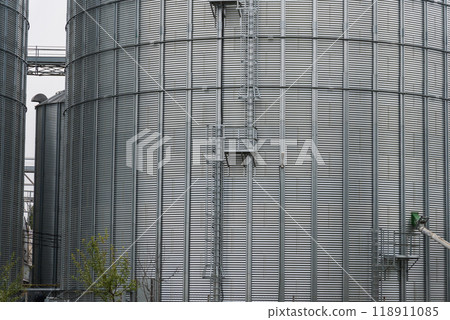 Tall metallic silos rise against the cloudy sky, showcasing access ladders and surrounding agricultural land. The structures store grains, vital for farming Tall metallic silos rise against the cloudy sky, showcasing access ladders and surrounding agricultural land. The structures store grains, vital for farming 118911085
