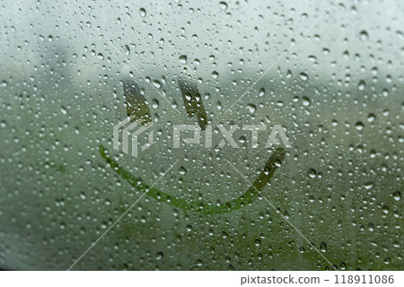 Raindrops create a textured surface on the glass, while a cheerful smiley face is drawn amidst the droplets, brightening the gloomy afternoon outside Raindrops create a textured surface on the glass, while a cheerful smiley face is drawn amidst the droplets, brightening the gloomy afternoon outside 118911086