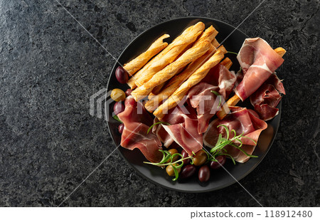 Prosciutto with bread sticks, olives, and rosemary on a black plate. 118912480