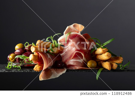 Prosciutto with bread sticks, olives, and rosemary on a black background. Prosciutto with bread sticks, olives, and rosemary on a black background. 118912486