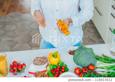 Female hands holding pill organizer with nutritional supplements on table with fresh food ingredients. Balanced Diet. Supplementing diet with missing vitamins. Dietary Daily intake for woman health 118913012