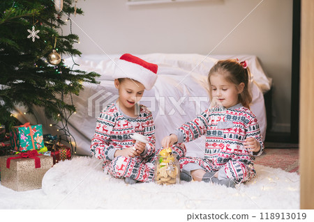 Children in festive pajamas excitedly explore a jar of holiday treats beside a decorated Christmas tree during the festive season. Joyful siblings sit on cozy rug, getting into jar filled with sweets 118913019