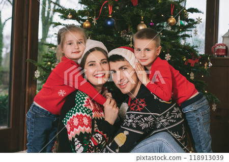 Smiling family portrait in festive sweaters sitting with gifts under Christmas tree at home during holiday. Happy parents and kids cuddling, feeling excited. Family time together. Happy childhood Smiling family portrait in festive sweaters sitting with gifts under Christmas tree at home during holiday. Happy parents and kids cuddling, feeling excited. Family time together. Happy childhood 118913039