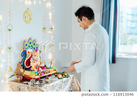Indian man dressed in traditional wear engages in worship at home altar, adorned with decors and offerings for god Ganesh during festive celebration. Indian culture, hindu ritual and customs. 118913046