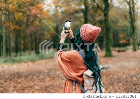 Woman wrapped in Shawl Poncho with backpack making photo by phone while enjoying autumn walk in forest. Capture the moment. Feeling harmony. Relaxing, personal fulfilment. Local travel lifestyle 118913061