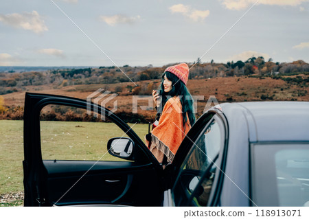 Hipster woman enjoying warm drink on car hood while taking in serene rural landscape during cold season road trip. Female wrapped in Shawl Poncho Cape and hat. Fall, winter travel, vacation concept. Hipster woman enjoying warm drink on car hood while taking in serene rural landscape during cold season road trip. Female wrapped in Shawl Poncho Cape and hat. Fall, winter travel, vacation concept. 118913071