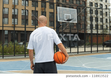 Basketball player with orange basketball ball on sports court courtyard. Basketball Hoop. View from back. Urban public park outside. 118913522