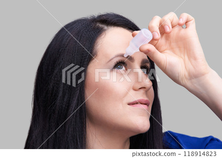 Woman applying eye drops against gray background for eye hydration and relief 118914823