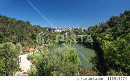 Landscape with a path in a sand park without people among trees and bushes and a green lake Landscape with a path in a sand park without people among trees and bushes and a green lake 118915374