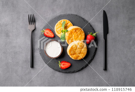 Cottage cheese, cheesecakes or syrniki with sour cream, strawberries and mint on a black slate Cottage cheese, cheesecakes or syrniki with sour cream, strawberries and mint on a black slate 118915746