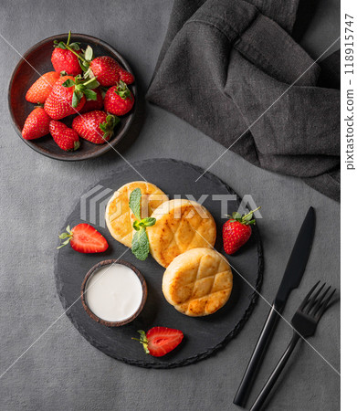 Cottage cheese, cheesecakes or syrniki with sour cream, strawberries and mint leaf on a black slate Cottage cheese, cheesecakes or syrniki with sour cream, strawberries and mint leaf on a black slate 118915747