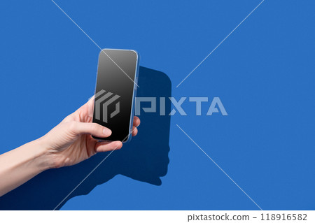 A hand holding a smartphone with a blank screen, with a harsh shadow on a blue background. 118916582