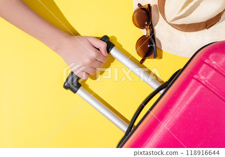 A hand holds a pink suitcase with travel accessories on a yellow background. 118916644