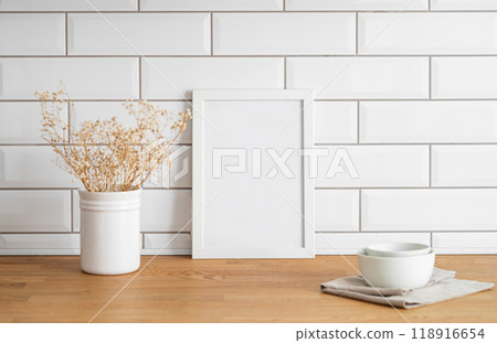 A frame with a blank canvas against a white tile wall and on a wooden tabletop with vase and cup. A frame with a blank canvas against a white tile wall and on a wooden tabletop with vase and cup. 118916654
