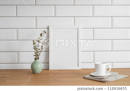 A frame with a blank canvas against a white tile wall and on a wooden tabletop with vase and cup. A frame with a blank canvas against a white tile wall and on a wooden tabletop with vase and cup. 118916655