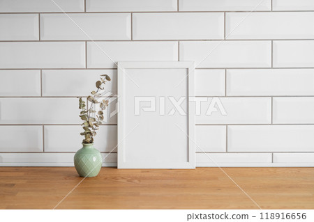A frame with a blank canvas against a white tile wall and on a wooden tabletop with vase. A frame with a blank canvas against a white tile wall and on a wooden tabletop with vase. 118916656