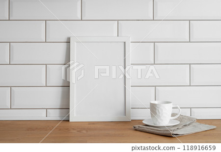 A  frame with a blank canvas against a white tile wall and on a wooden tabletop with cup of tea. 118916659