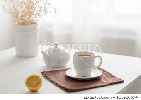 A cup of tea with lemon and a teapot on a white table with dry bouquet against the background  118916674