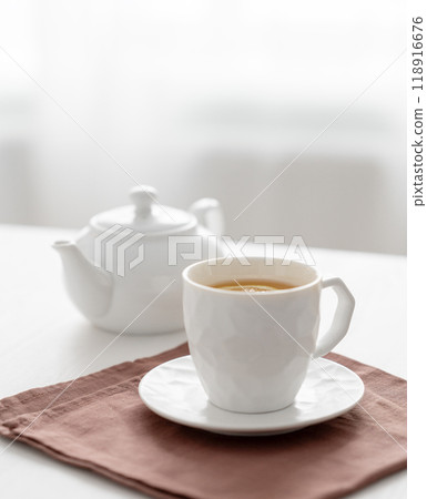 A cup of tea with lemon and a teapot on a white table against the background of a kitchen window. A cup of tea with lemon and a teapot on a white table against the background of a kitchen window. 118916676