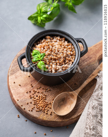 Buckwheat porridge in a black pan on a cutting board on a light background with raw grains, spoon Buckwheat porridge in a black pan on a cutting board on a light background with raw grains, spoon 118916815