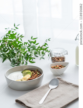 Granola with banana and kiwi on a white table with a green branch against the background of a window Granola with banana and kiwi on a white table with a green branch against the background of a window 118916862