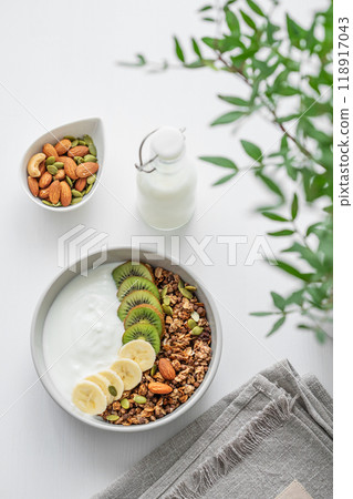 Granola with banana and kiwi on a white table with a green branch. 118917043
