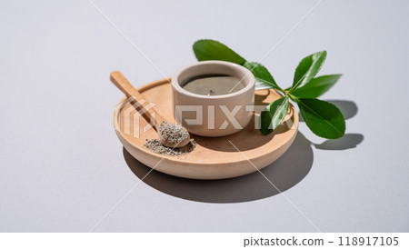 Blue clay in a bowl and spoon on a wooden plate on a light background with leaves and shadow. 118917105