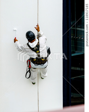 A worker paints the hull of a white ship with a paint roller while hanging from a rope A worker paints the hull of a white ship with a paint roller while hanging from a rope 118917185
