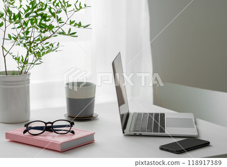 Desktop with laptop, coffee cup, phone, notepad and glasses in front of a window Desktop with laptop, coffee cup, phone, notepad and glasses in front of a window 118917389