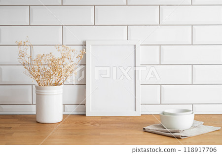 A  frame with a blank canvas against a white tile wall and on a wooden tabletop with vase and cup. 118917706