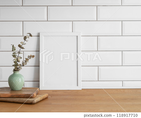 A  frame with a blank canvas against a white tile wall and on a wooden tabletop with vase. 118917710